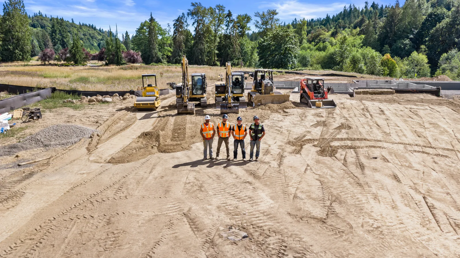 	Excavation team photo on a leveled building pad with a roller, two excavators, dozer, and track loader lined up behind them