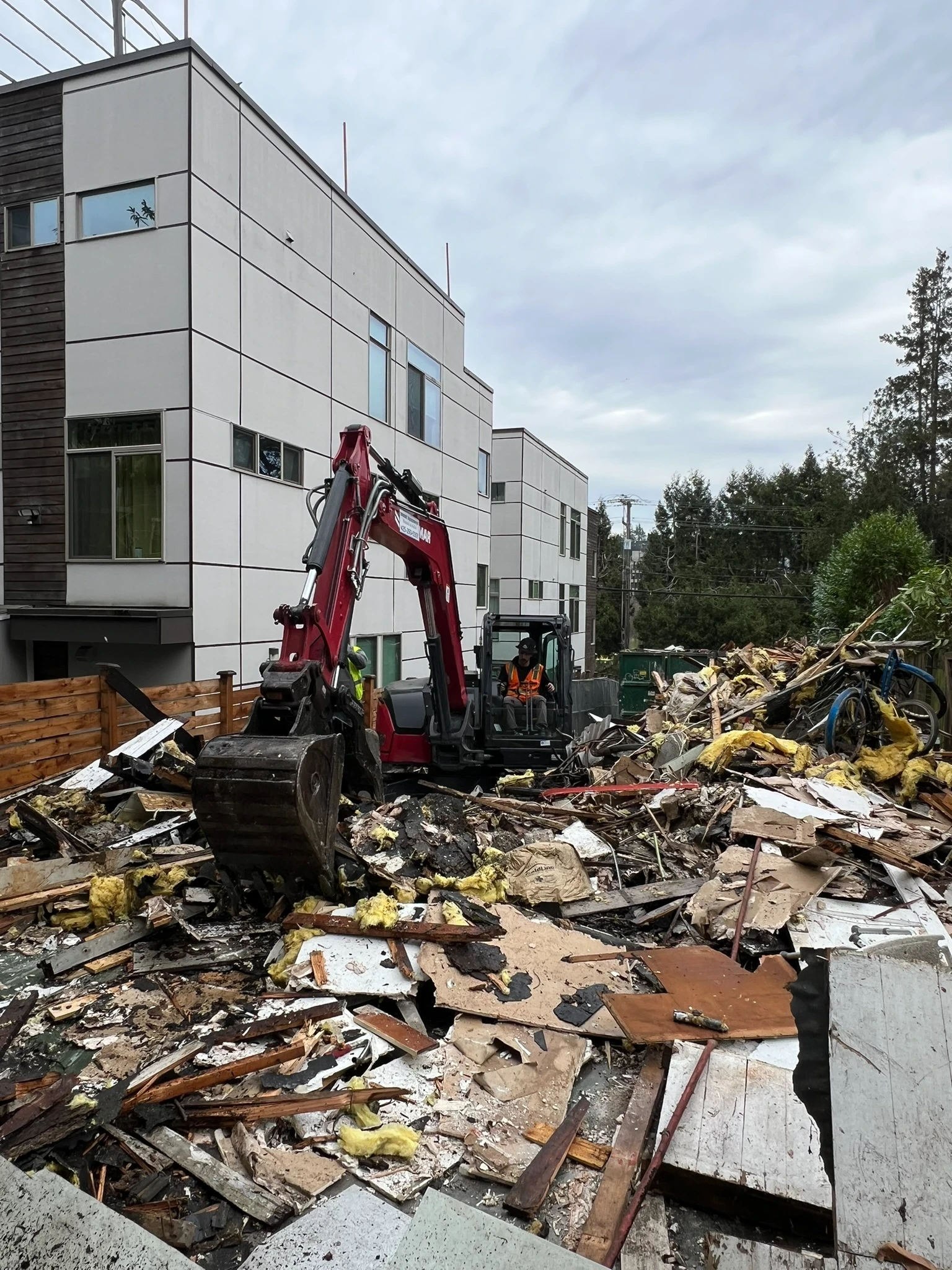Red excavator clearing debris and wooden rubble during a building demolition near modern townhomes. 