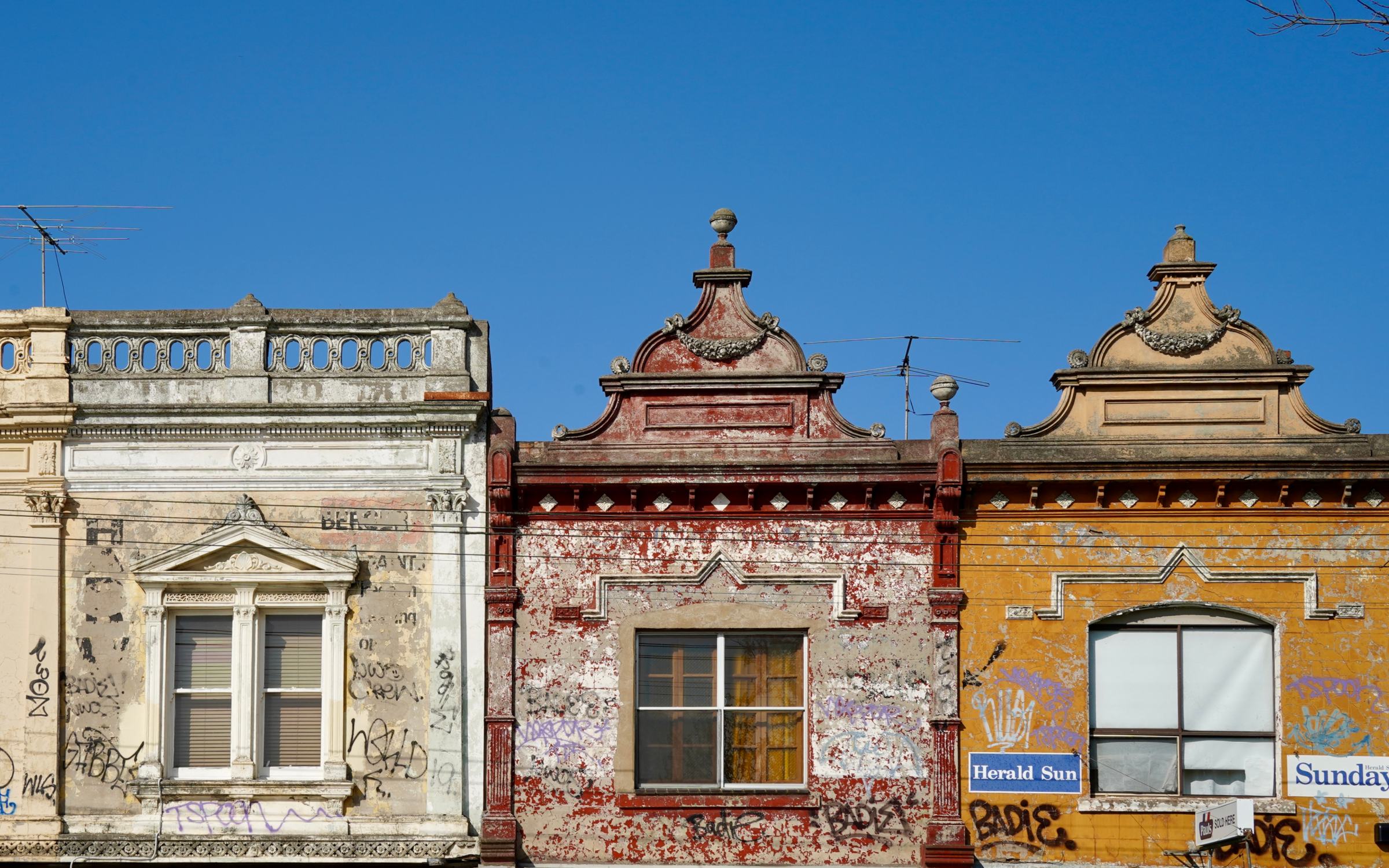 Three heritage shopfront parapets in cream, red brick and ochre against a blue sky, Melbourne's inner north