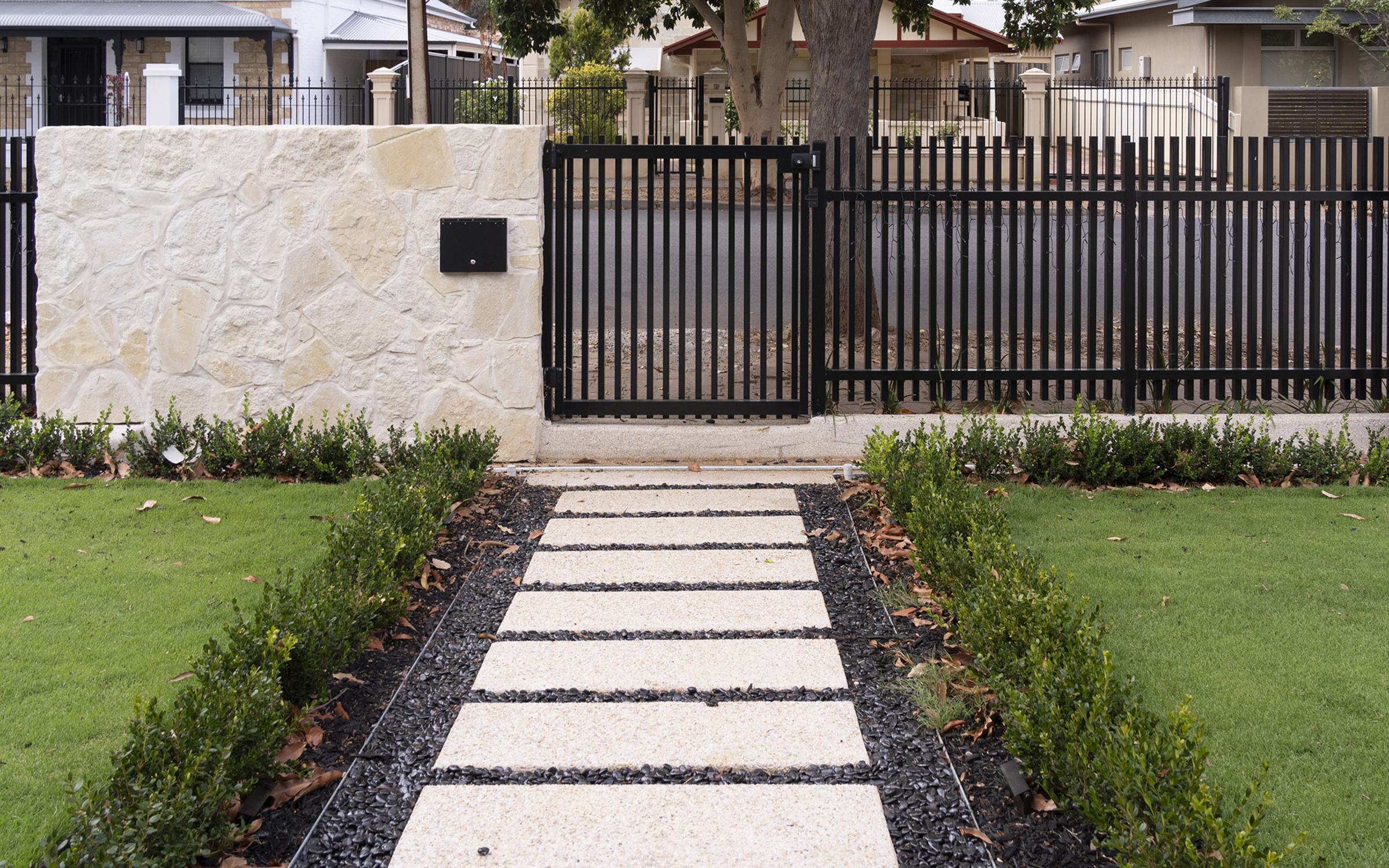 Exposed aggregate stepping-stone path through a lawn to a sandstone wall and black steel gate, built by Catcrete