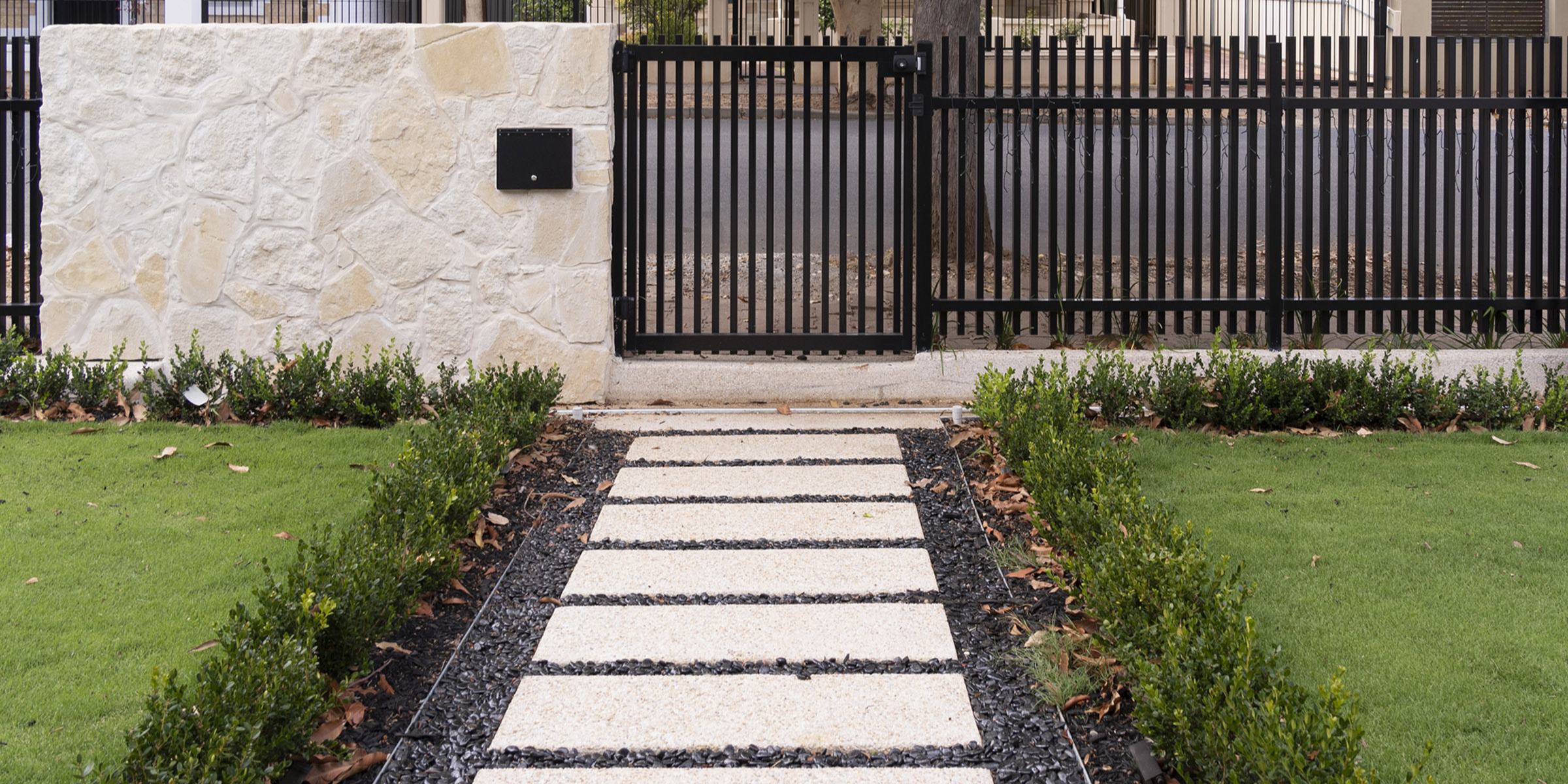 Exposed aggregate stepping-stone path through a lawn to a sandstone wall and black steel gate, built by Catcrete