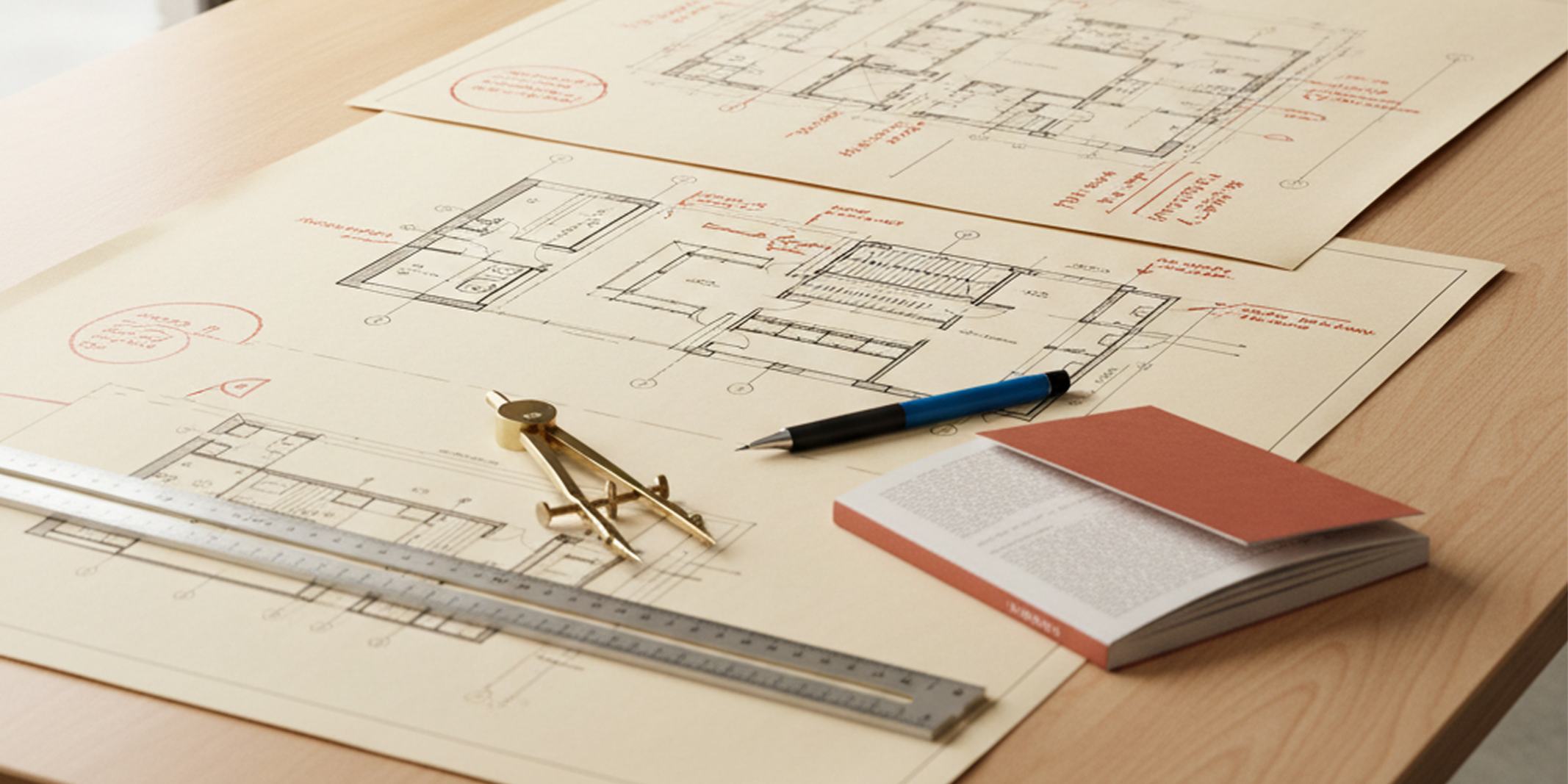 Architectural plans on a timber desk with a brass compass, steel rule, pen and notebook