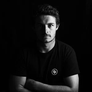 Smiling man with short hair and a mustache wearing a black polo shirt with a small green logo on the chest, standing in front of a blurred white background with green text.