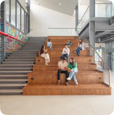 People sitting and talking on wooden tiered seating in a modern indoor space with stairs and colorful wall art.