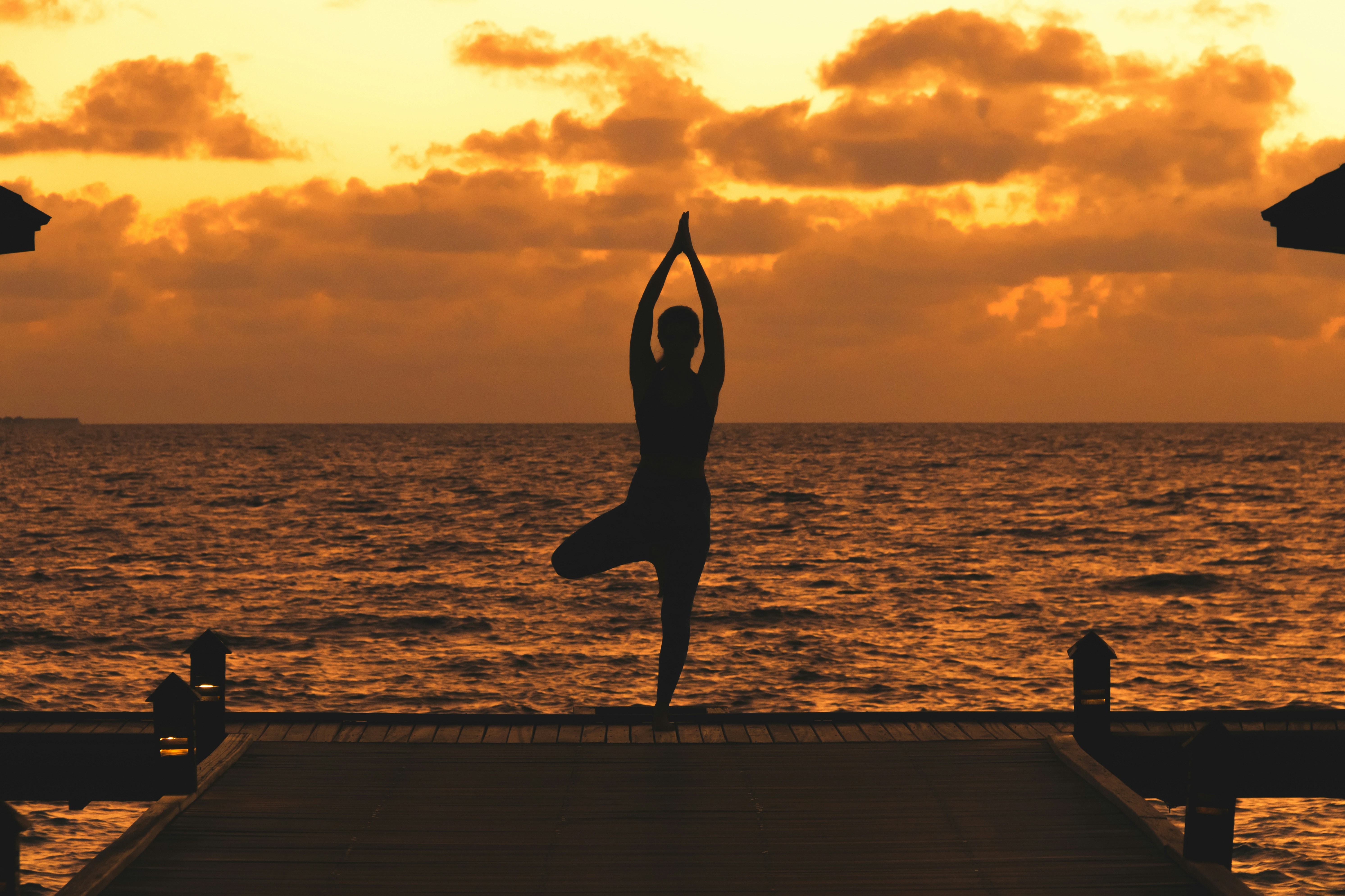 Silhouette of a person doing a yoga tree pose on a dock at sunset over the ocean.