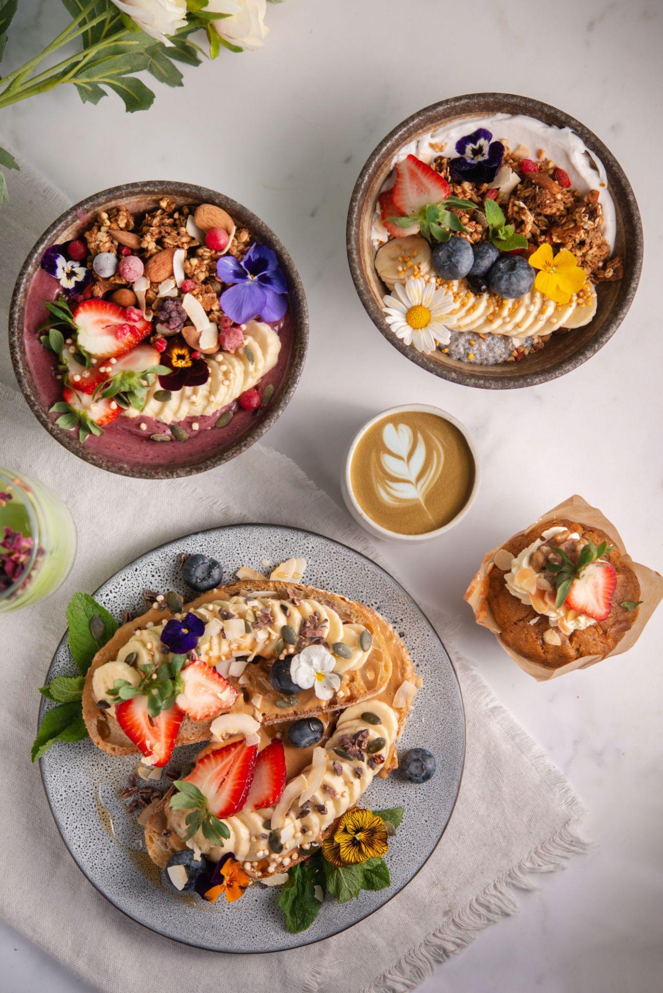 Plate with banana toast and berries, smoothie bowls with fruit and nuts, muffin, and latte with leaf art.