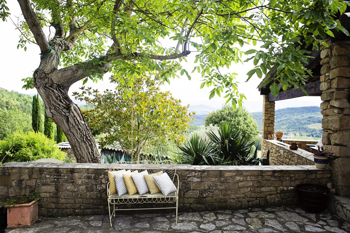 Outdoor stone patio with a metal bench and patterned cushions overlooking green trees and hills.
