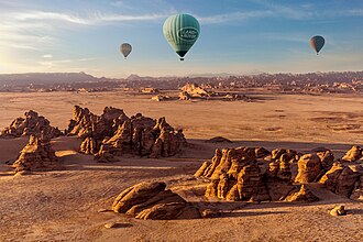 Three colorful hot air balloons flying over rocky desert landscape with mountains under blue sky.