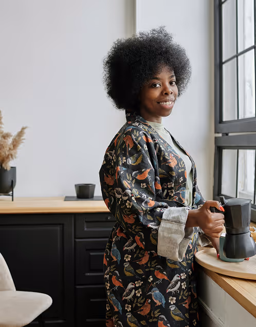 Smiling woman with curly hair wearing a patterned robe holding a stovetop coffee maker by a kitchen window.