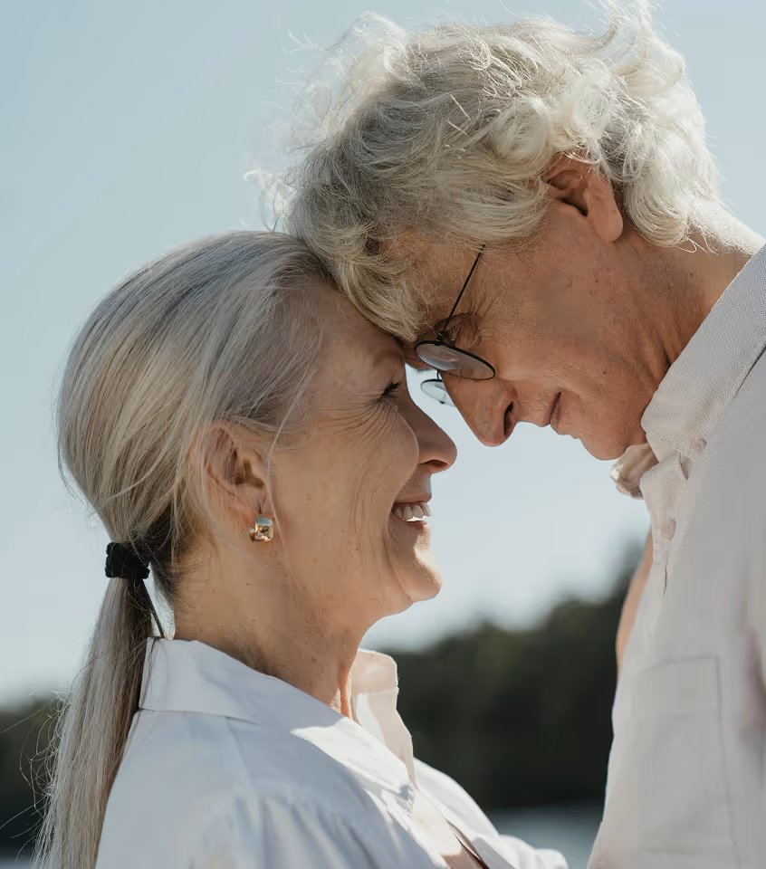 Smiling elderly couple touching foreheads outdoors under clear sky.