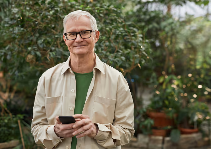 Smiling elderly man with glasses holding a smartphone, standing outdoors with greenery in the background.