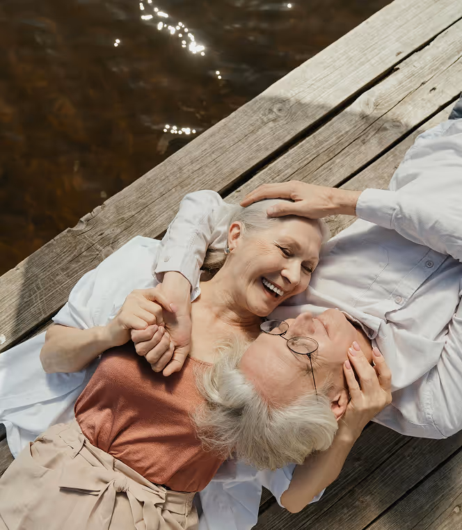 Elderly couple lying on a wooden dock by the water, smiling and holding each other affectionately.