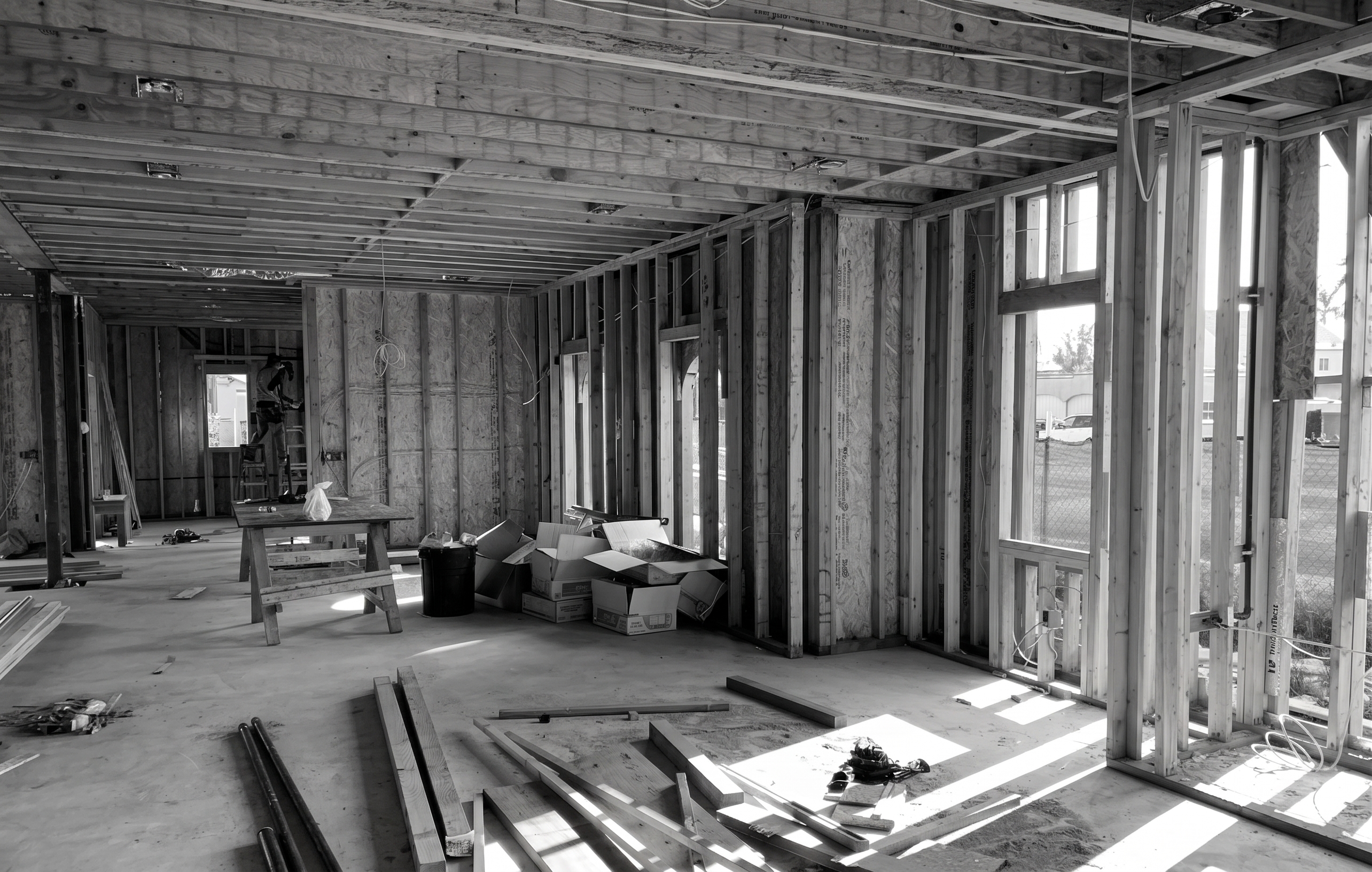image of a house interior during the framing stage of construction, featuring exposed wooden wall studs, ceiling joists, and scattered lumber on a concrete floor.