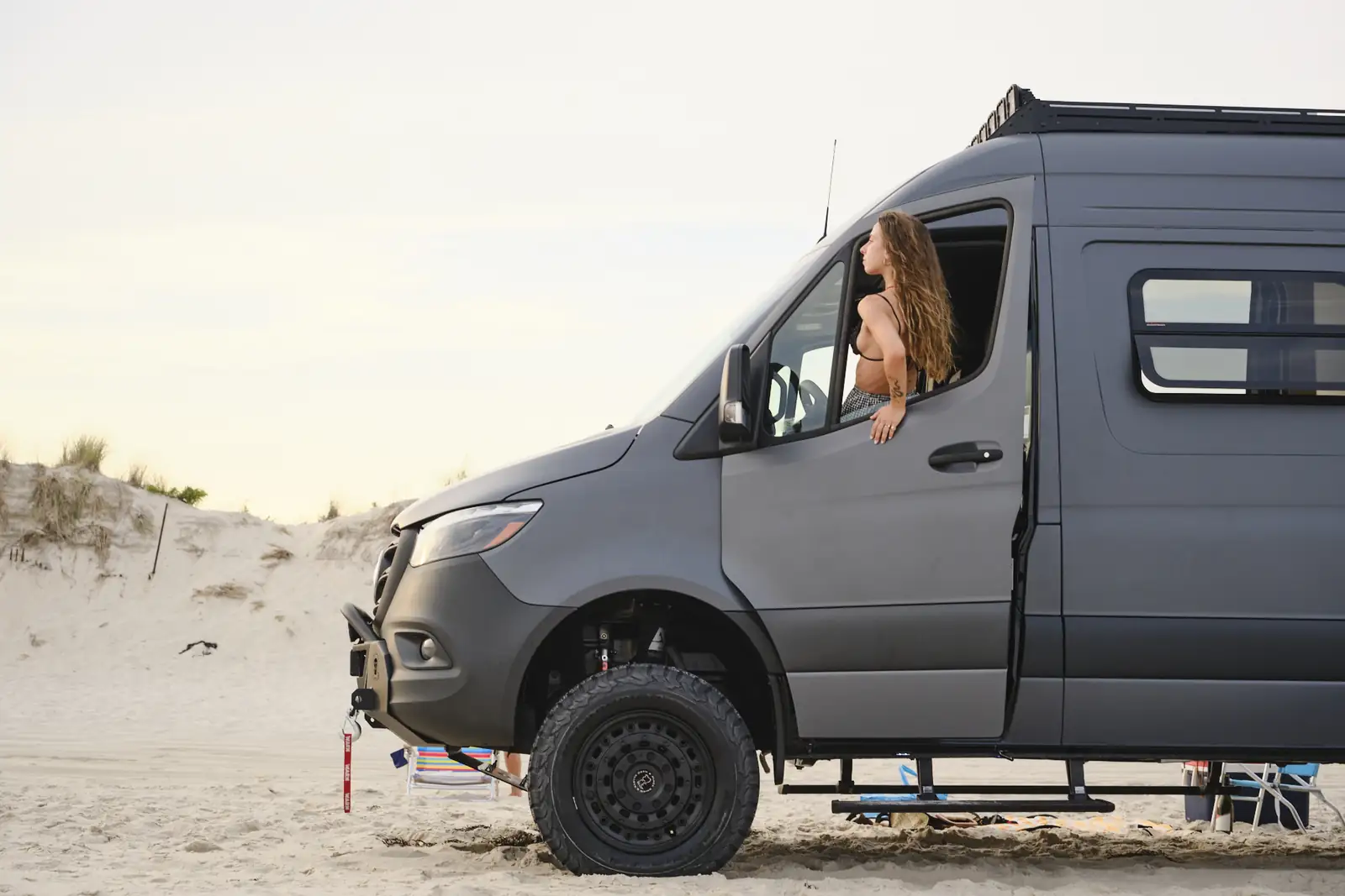 Grey 4x4 Brooklyn Campervans Mercedes Sprinter on the beach with a woman leaning out of the window