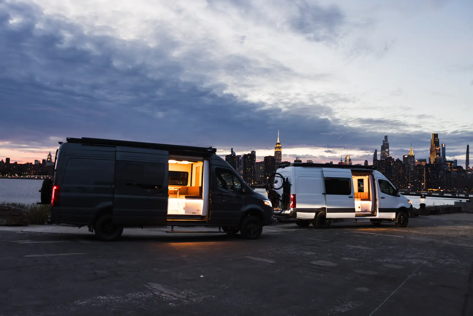 Two Mercedes Sprinter campers on a Brooklyn pier at dusk with lit interiors and the Manhattan skyline behind