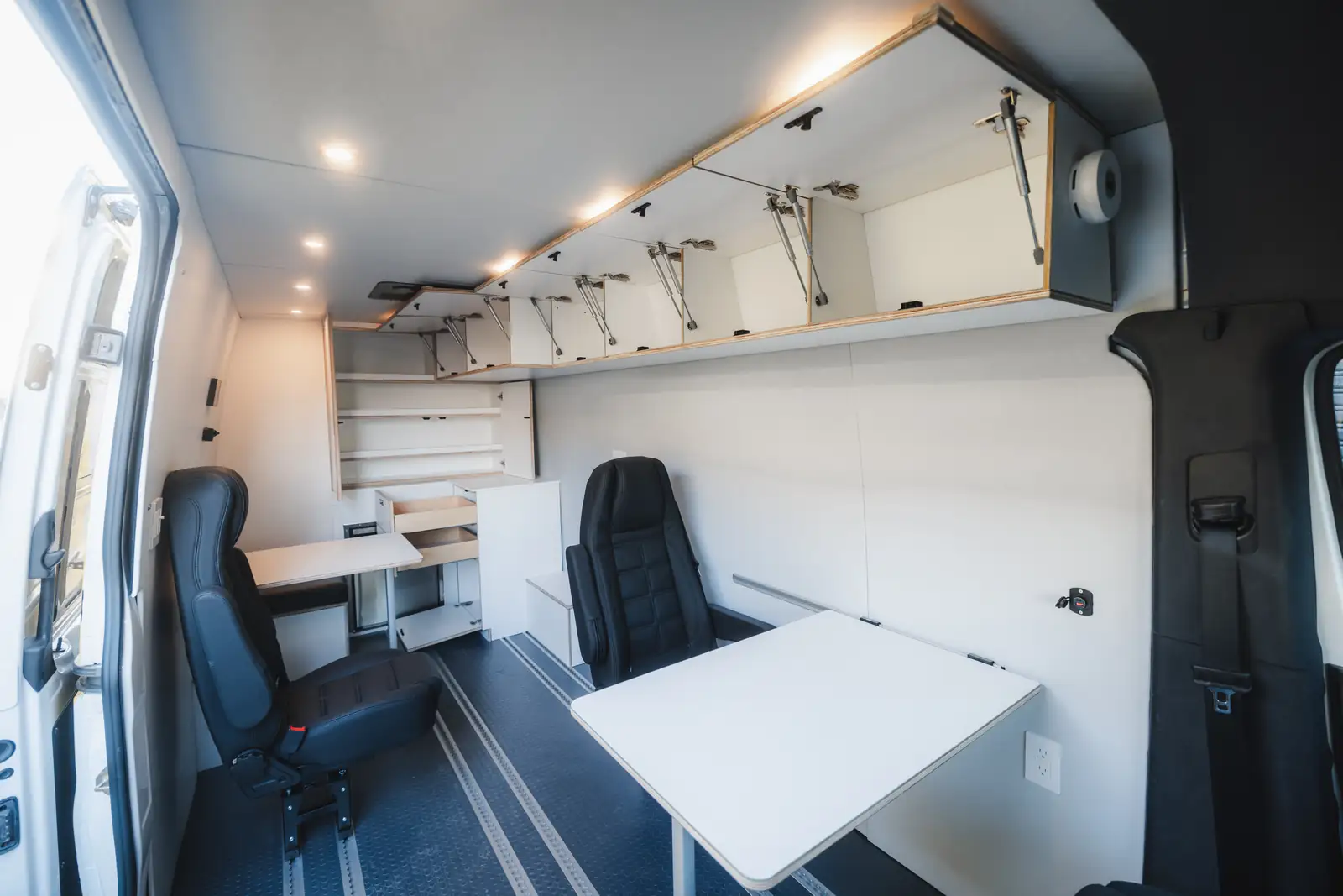 Sprinter office interior with two black office chairs, a fold-down white table and open overhead cabinets