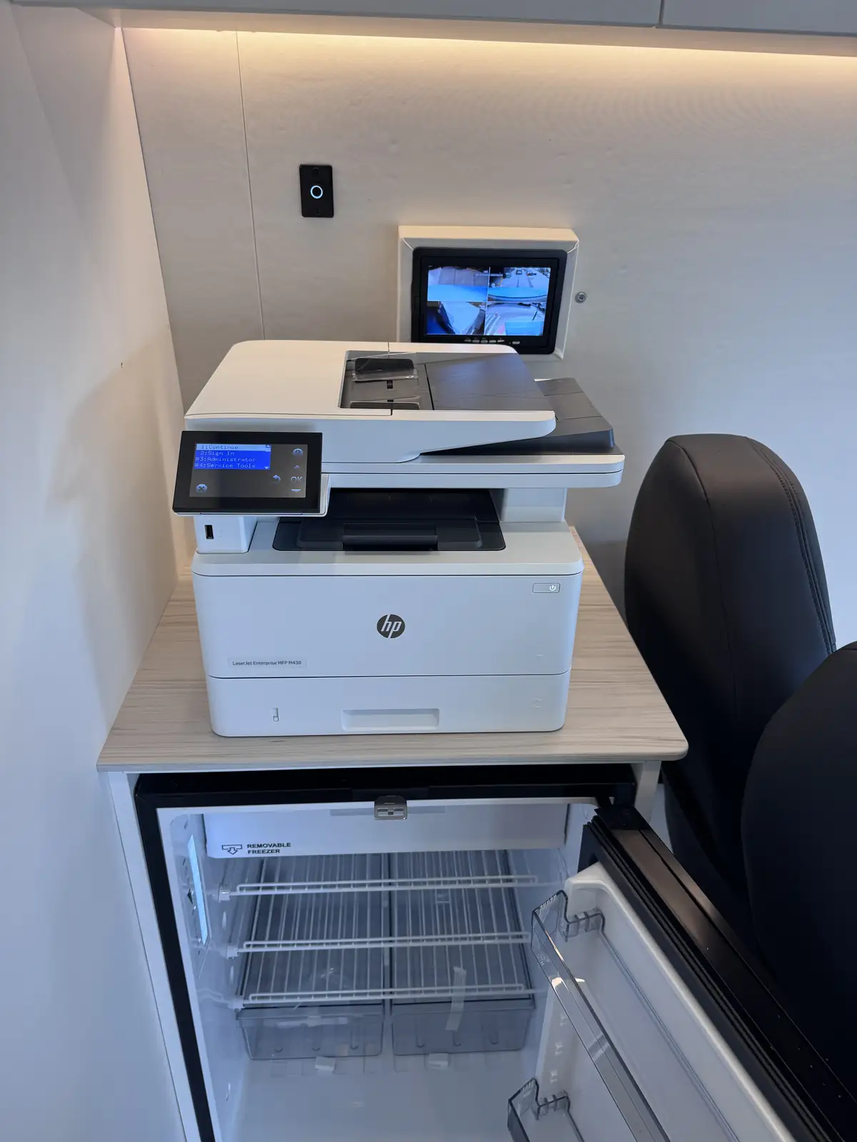 Multifunction printer on a wood counter above an open under-counter fridge, with a camera monitor on the wall