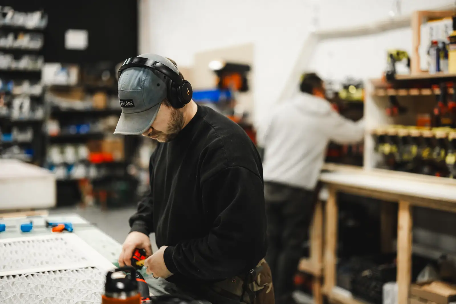 Two builders at work in the shop, one measuring at a workbench beside white panels and another at the tool wall