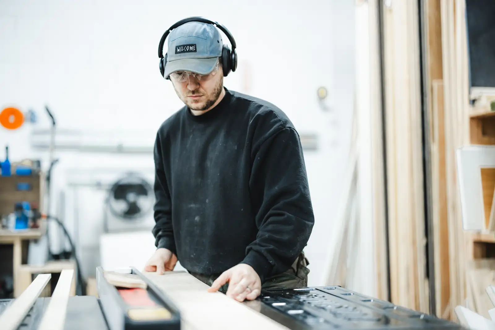 Builder in safety glasses guiding a board across a table saw, with a lumber rack beside him