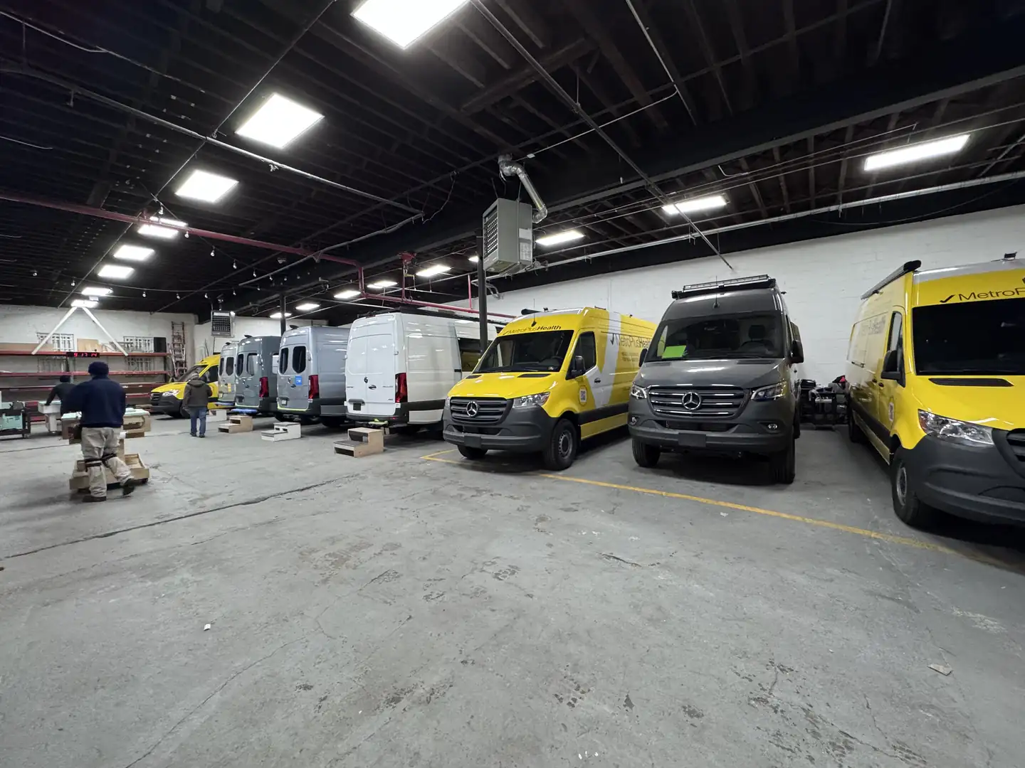 Workshop floor with several Mercedes Sprinters including two yellow MetroPlus vans and two workers loading boxes