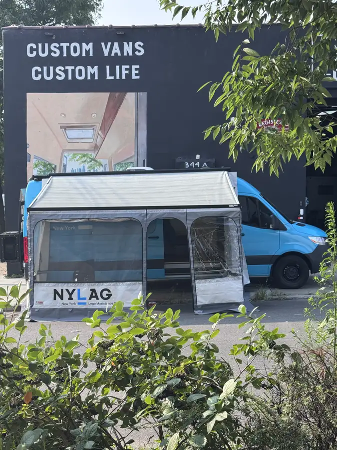 NYLAG New York Legal Assistance Group light blue Mercedes Sprinter with side awning and tent set up outside the shop