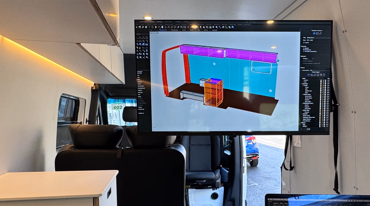 Wall-mounted TV inside a van showing a 3D layout model of the build, with white cabinets and LED strip lighting