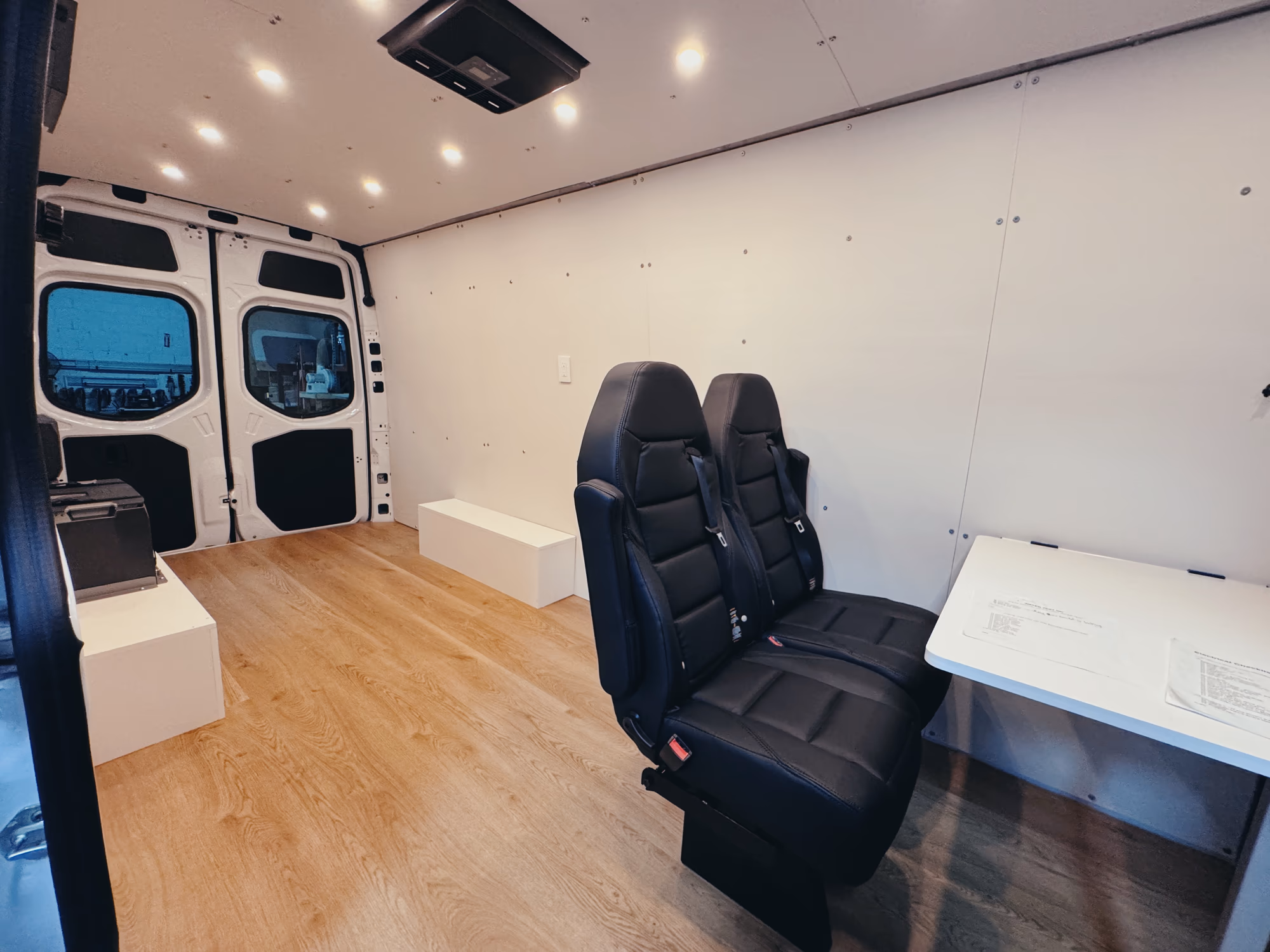 Empty Sprinter van interior with wood floor, two black leather seats, a fold-down white desk and ceiling lights