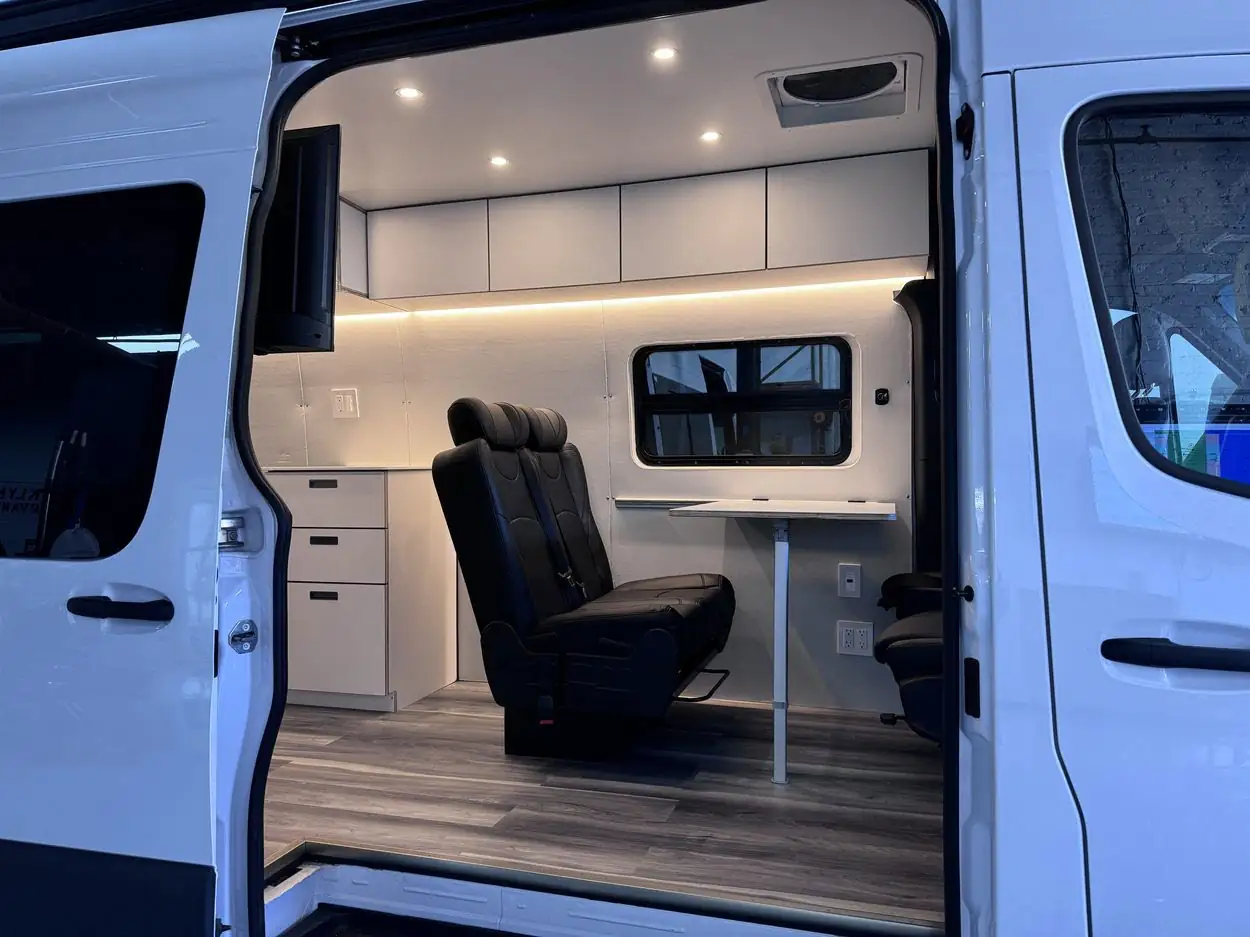 View through the open sliding door of a white Mercedes Sprinter to a white drawer cabinet, black leather bench and table