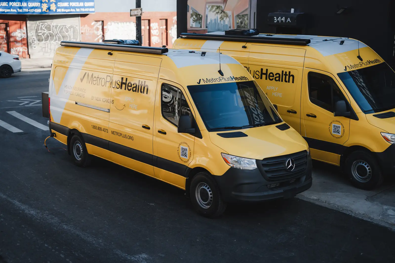Two yellow MetroPlusHealth Mercedes Sprinter vans with roof awnings parked on a Brooklyn street