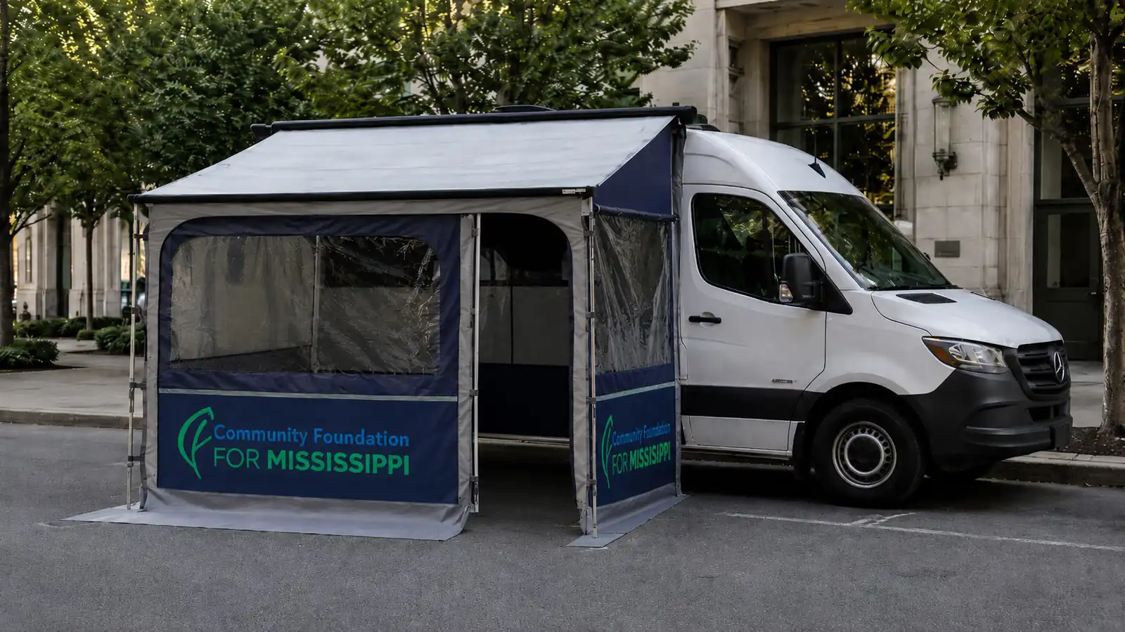White Mercedes Sprinter with side awning and Community Foundation for Mississippi tent outside a stone building at dusk