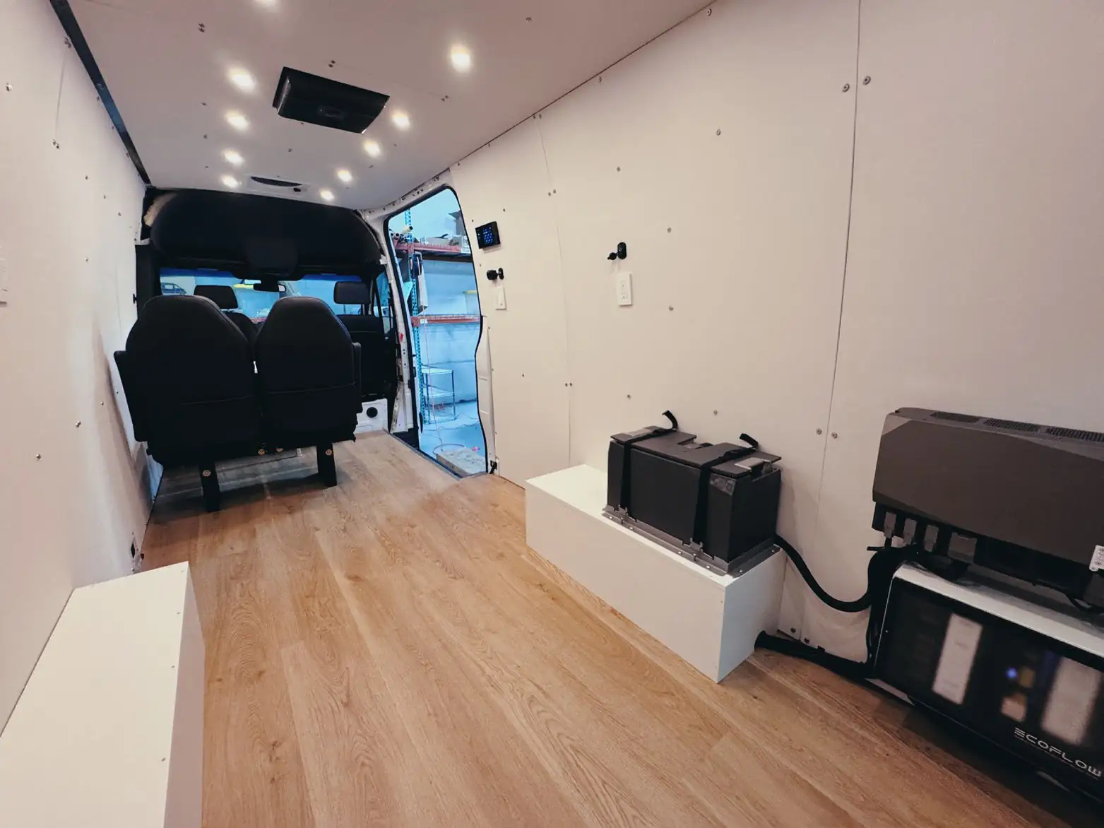 Nearly empty Mercedes Sprinter interior with white walls, light wood floor, two black seats and an EcoFlow battery bank