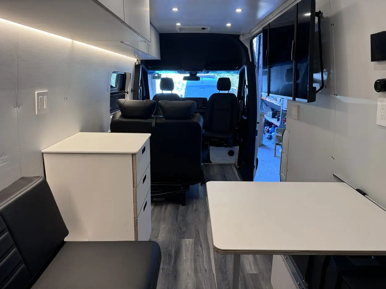 Mercedes Sprinter mobile office interior with white drawer cabinet, black leather bench, folding table and two wall TVs