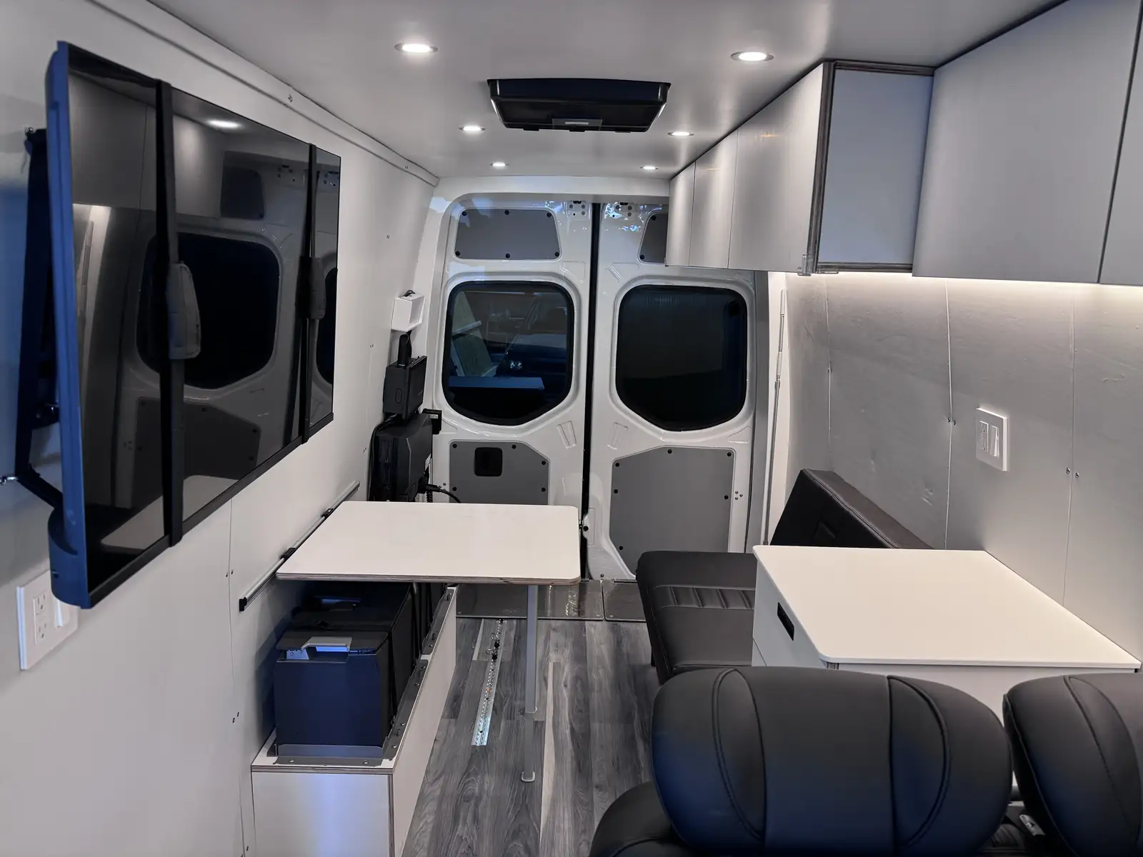 Van office interior with large wall-mounted TV, two white folding tables, black leather seats and grey overhead cabinets