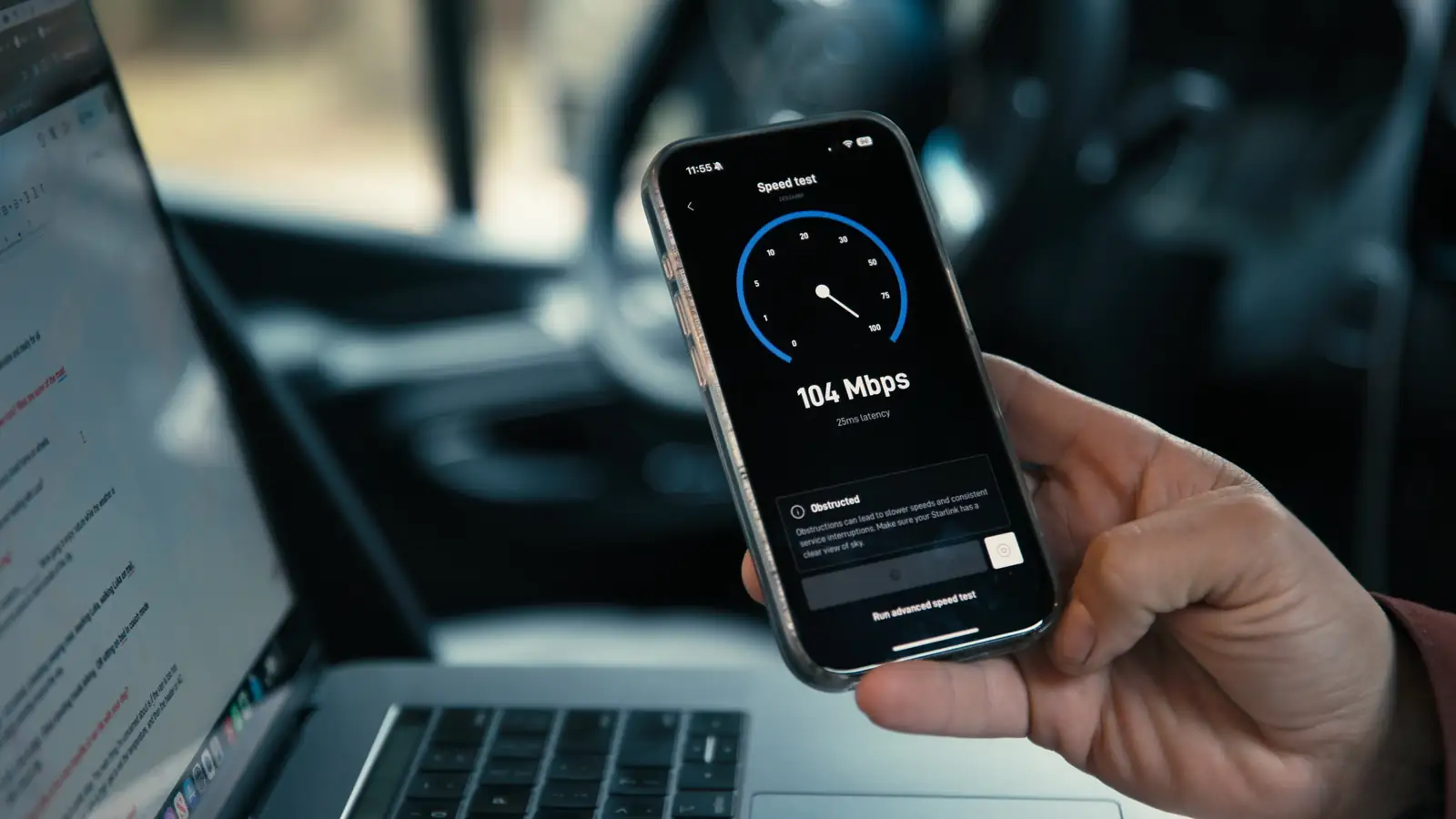 Hand holding an iPhone showing a 104 Mbps speed test next to an open laptop inside a vehicle