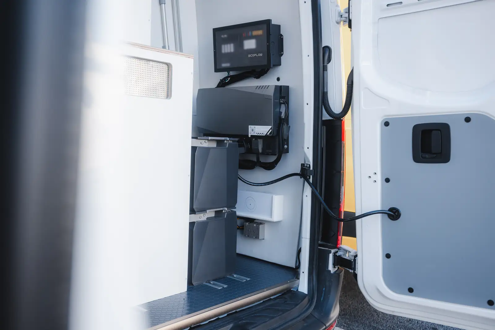 View through the open rear door of a white van to an EcoFlow screen, grey inverter and two black batteries