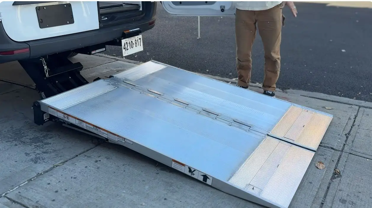 Aluminum lift deployed from the rear door of a white van down to the sidewalk, with a person standing beside it