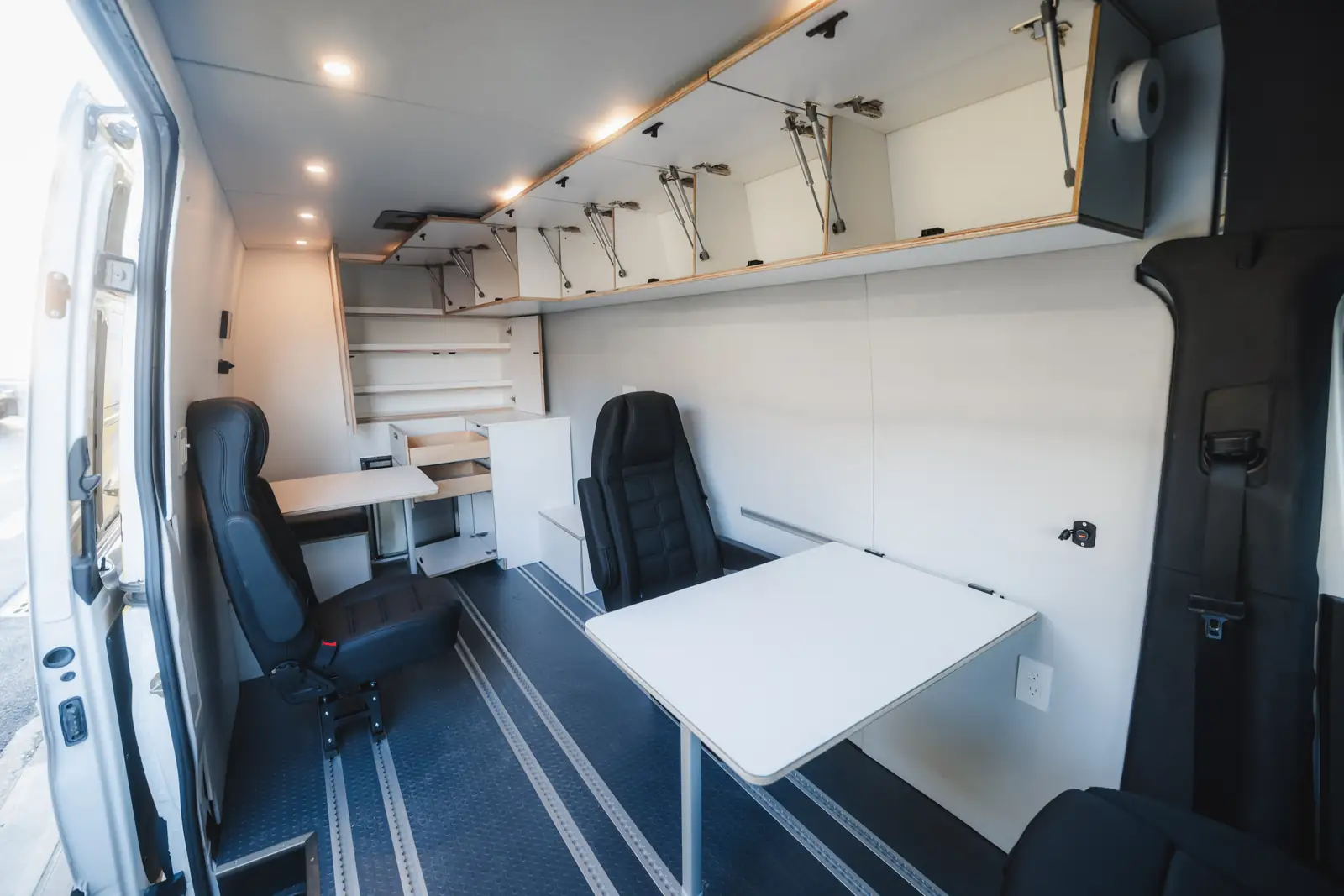 Mercedes Sprinter office interior from the side door with open overhead cabinets, two black swivel seats and blue floor