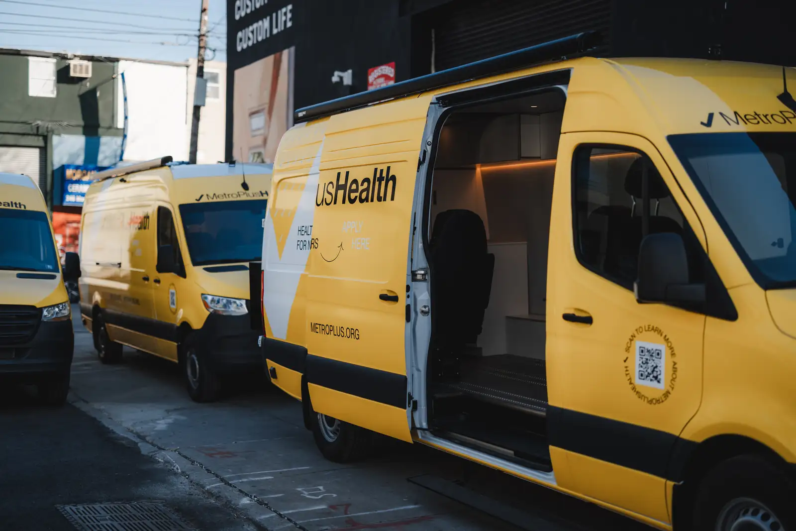 Yellow MetroPlusHealth Mercedes Sprinter with sliding door open showing a white interior, two more vans behind