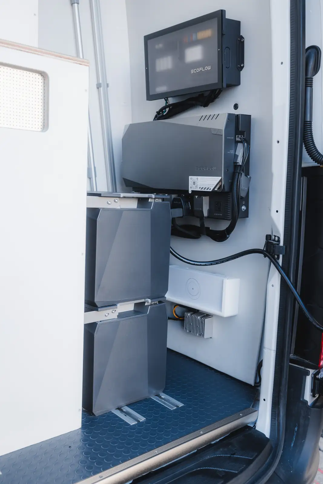 Rear corner of a white van with EcoFlow control screen, grey inverter and two stacked batteries on blue non-slip floor