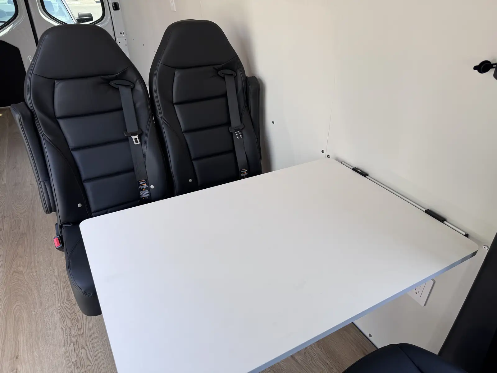 Two black leather seats with seat belts facing a white wall-mounted folding table on oak-look vinyl flooring