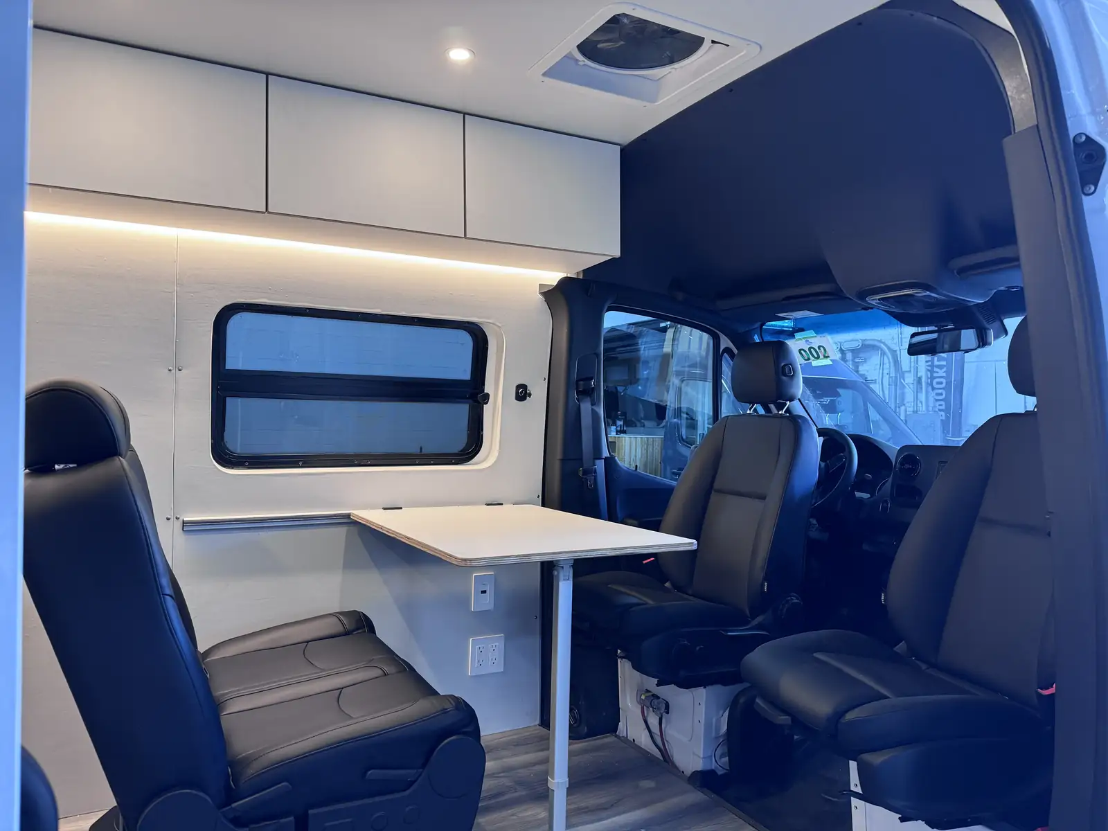 Van interior with light grey LED-lit overhead cabinets, black leather seat, white pedestal folding table and roof fan