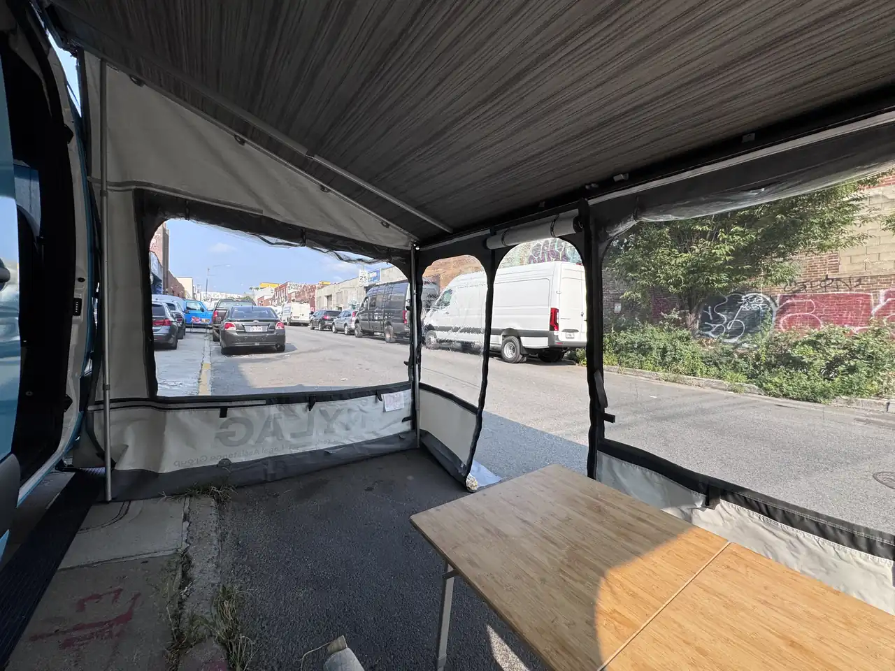 Side awning tent of a light blue Mercedes Sprinter with bamboo folding table, NYLAG print and the street beyond
