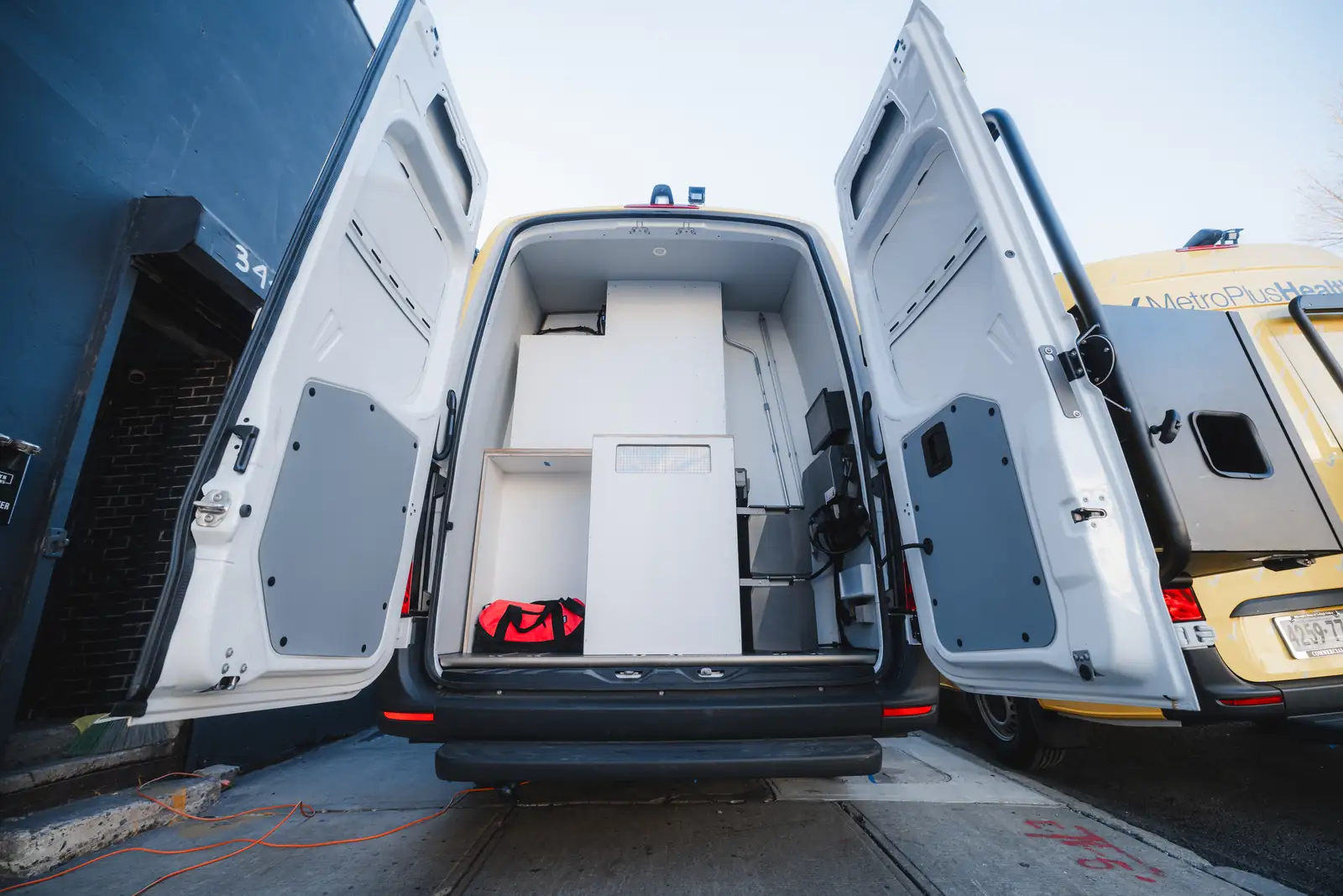 White Mercedes Sprinter with rear doors open showing white cabinetry, parked beside a yellow MetroPlusHealth vehicle