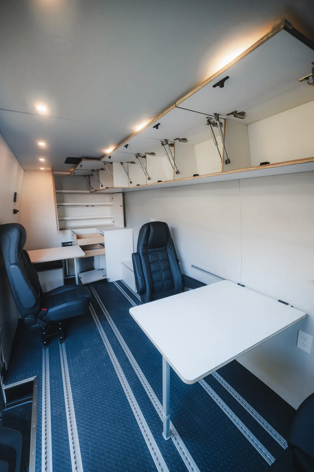 Van office interior with blue rubber floor, two black office chairs, large white pedestal table and open overhead cabinets