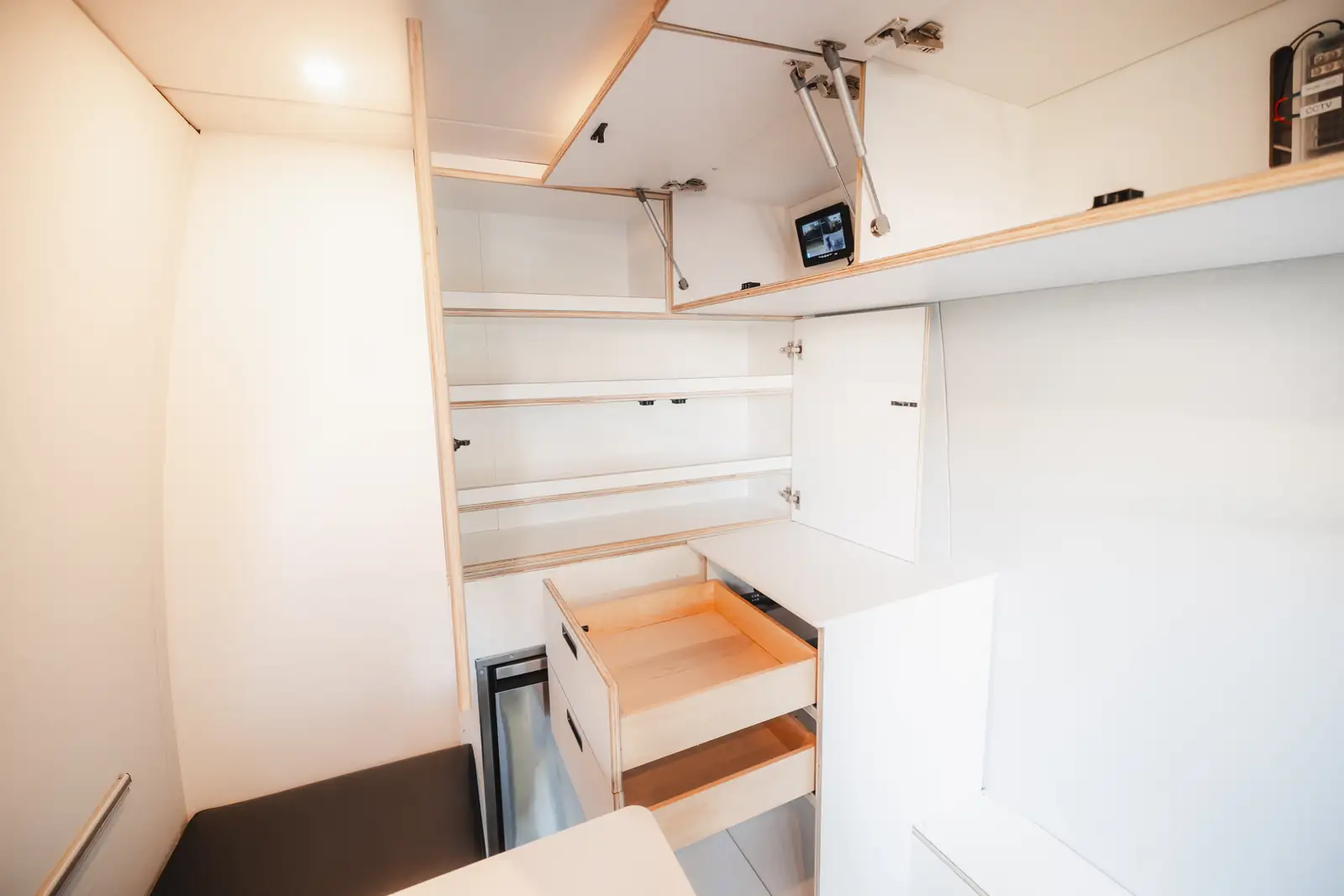 Van interior with white cabinets with light wood edges, open overhead doors on gas struts and two pulled-out drawers
