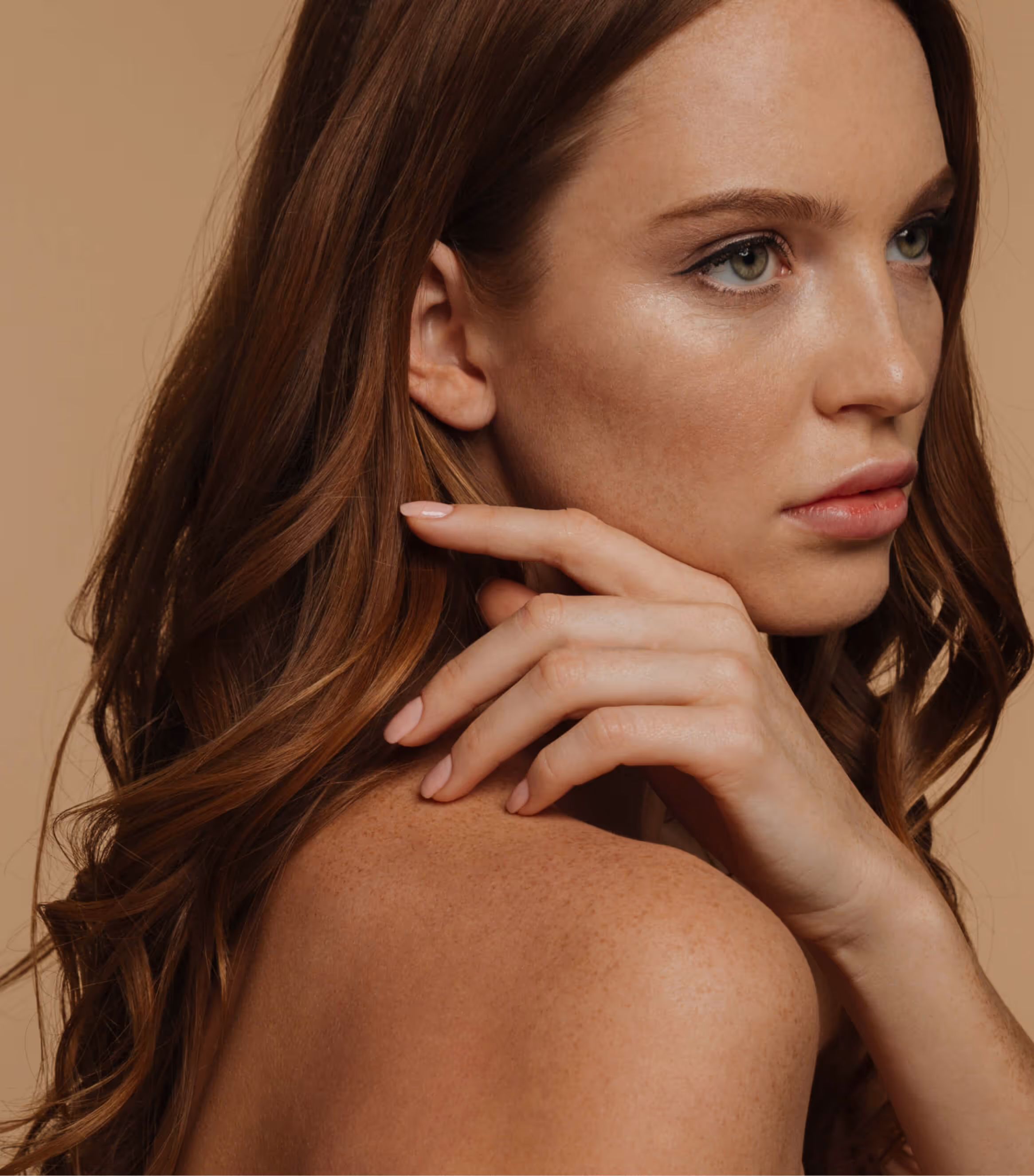 Close-up of a woman with long brown hair, green eyes, light makeup, and bare shoulders against a beige background.