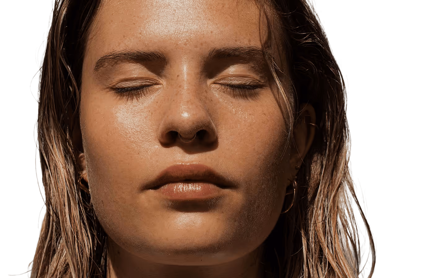 Close-up of a woman with closed eyes, wet blonde hair, and a septum ring under natural sunlight.