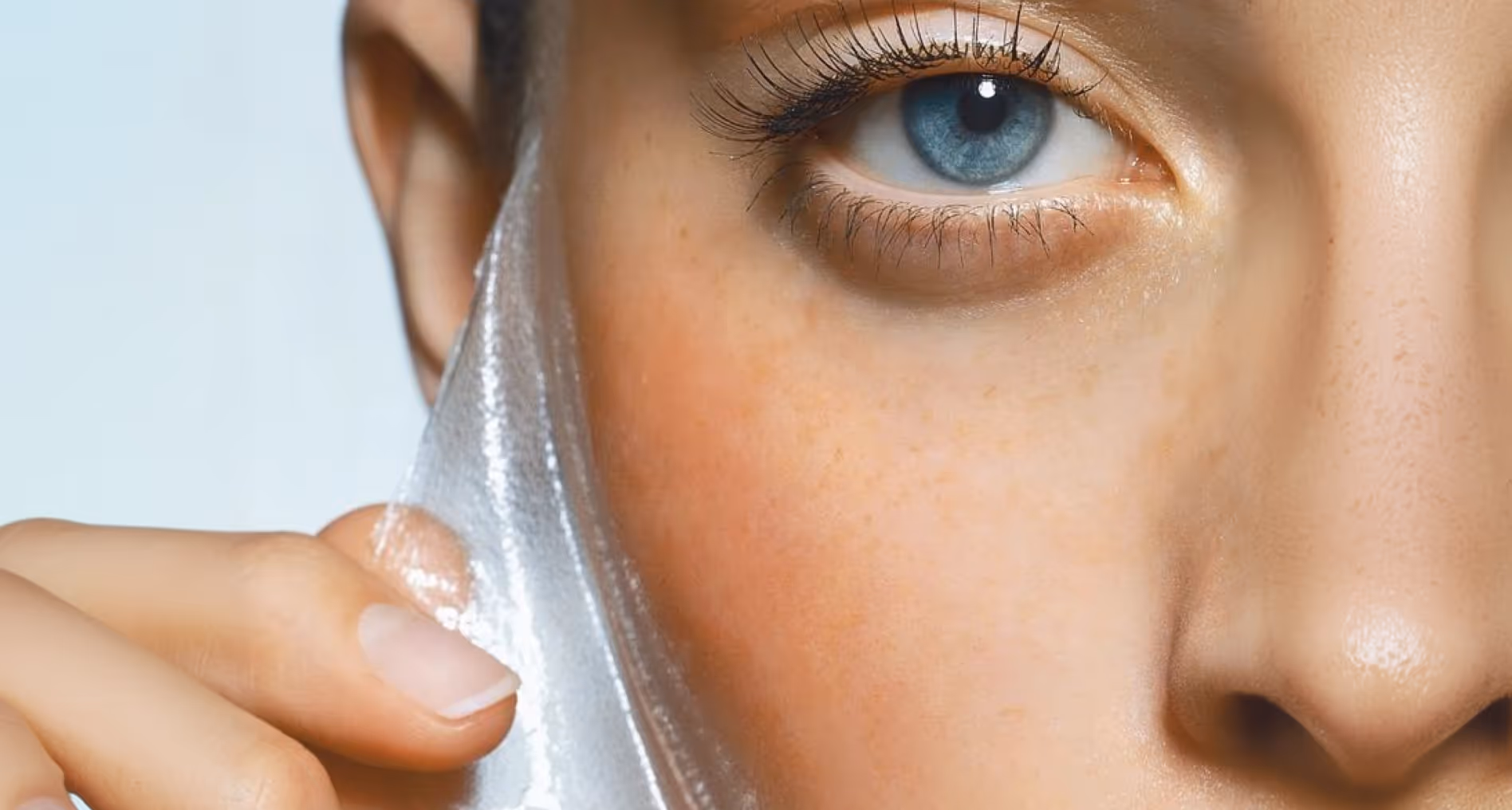 Close-up of a person peeling a transparent facial mask off their cheek near a blue eye.