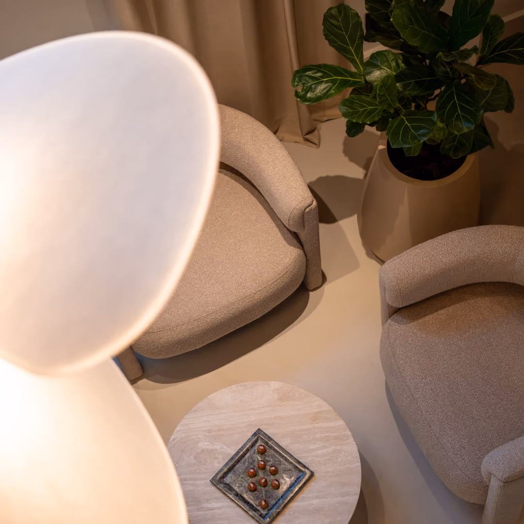 Modern living room with a curved brown sofa, two beige armchairs, a small round ottoman, two black side tables, a uniquely shaped coffee table, and a large vase with dried pampas grass.