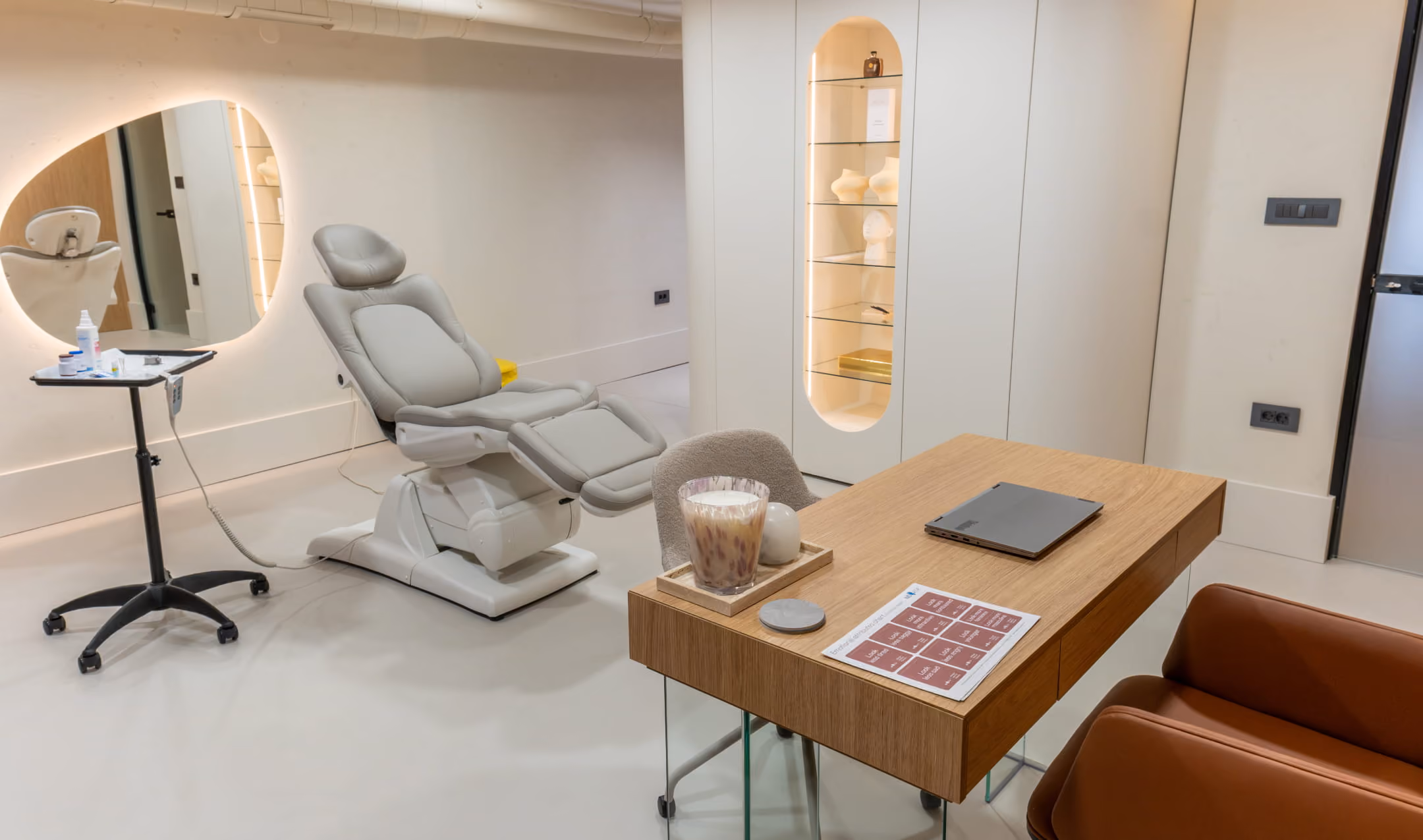 Modern, minimalistic beauty salon interior with beige treatment chair, stylish seating, a glass-topped coffee table, and a wooden desk with two chairs and decorative items.